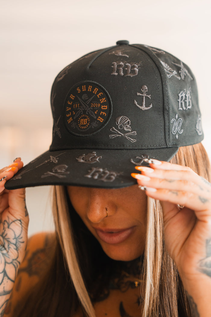 A person with long hair and tattooed hands holds the brim of a Relentless Betrayal Never Surrender Limited Edition Snapback, adorned with anchors, skulls, and bold lettering. Their nails are long, painted orange and white.