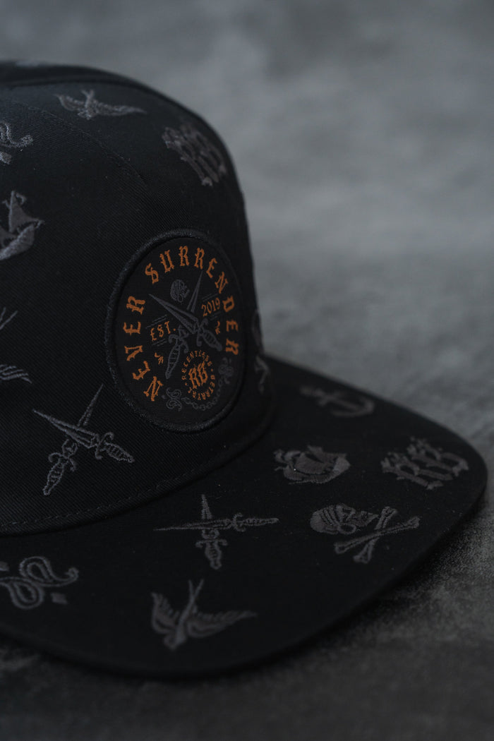 The Never Surrender Limited Edition Snapback by Relentless Betrayal is a black, handmade hat with tattoo-style embroidery and an orange "Never Surrender" patch, displayed on a dark textured surface.
