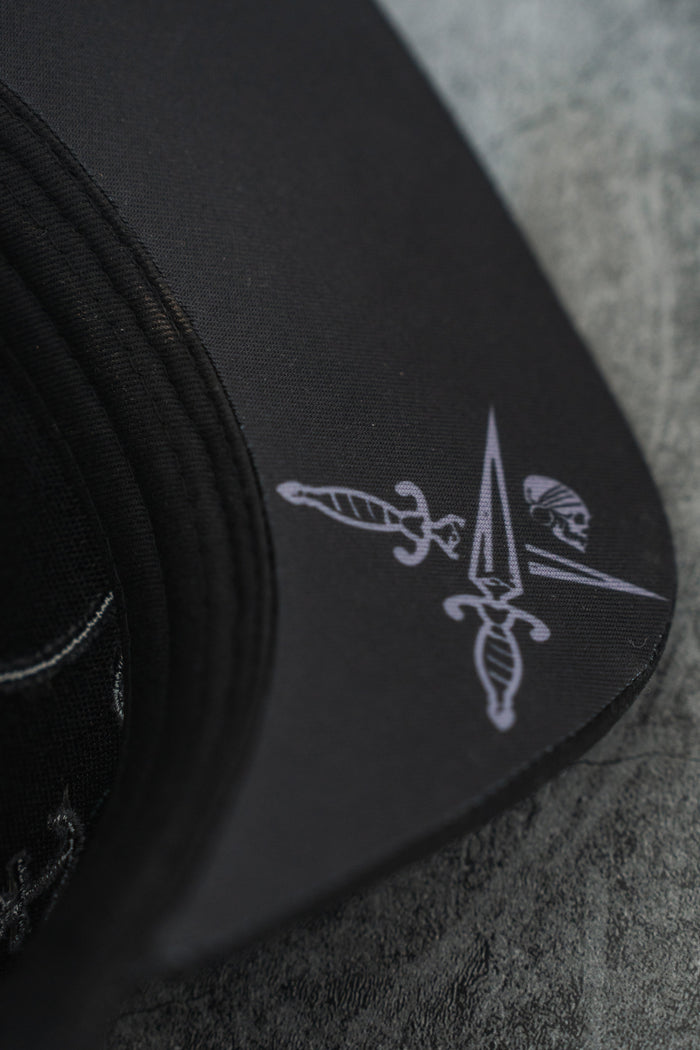 Close-up of the Never Surrender Limited Edition Snapback by Relentless Betrayal, featuring a printed design with two crossed daggers, a pointed spear, and a small skull on the brim, displayed on a grey textured surface.