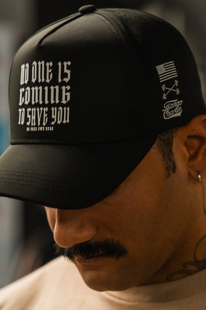 A tattooed person with a mustache looks down, wearing the Tango Charlie Apparel "No One Is Coming To Save You Hydro" snapback hat. The black cap features white gothic text, a small American flag, and additional side graphics.