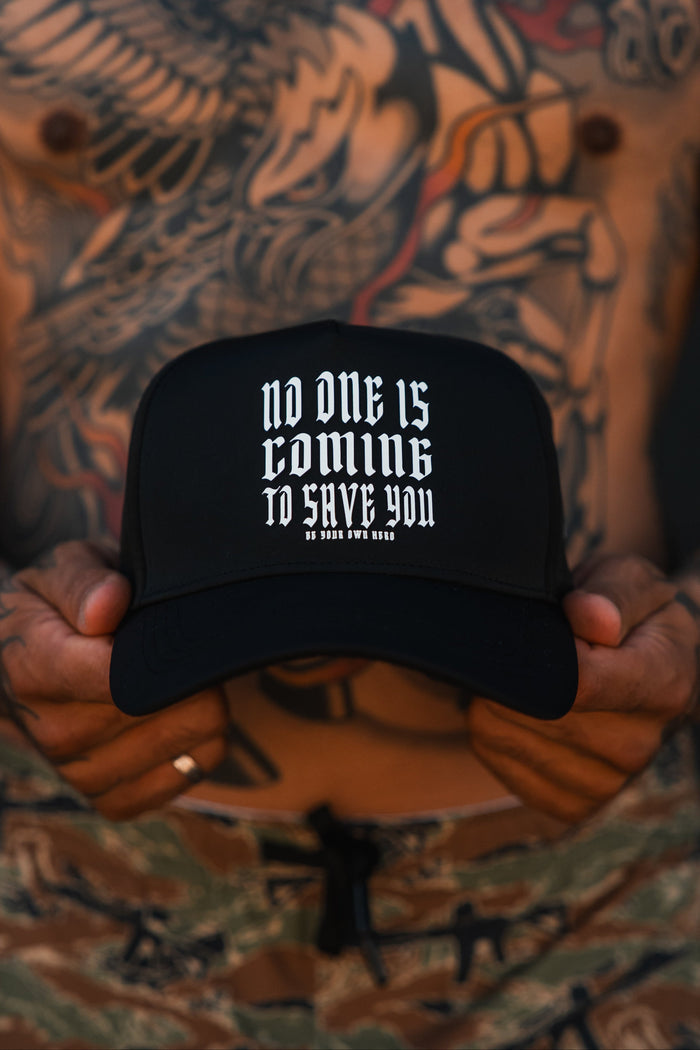 A tattooed person wearing camo pants holds the Tango Charlie Apparel "No One Is Coming To Save You Hydro - Snapback Hat," a black, hydro-resistant hat with bold white text. The blurred background highlights the hat and hands.