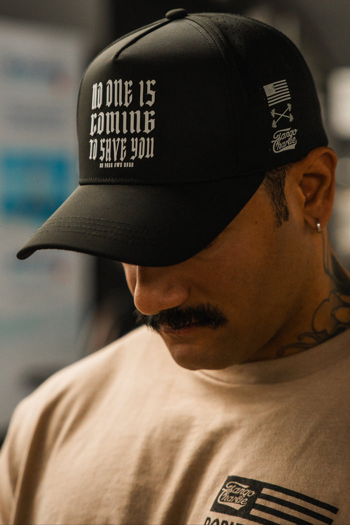 A person with a mustache and neck tattoos wears the Tango Charlie Apparel No One Is Coming To Save You Hydro-Snapback Hat in black, featuring white text, paired with a beige T-shirt with a small logo.
