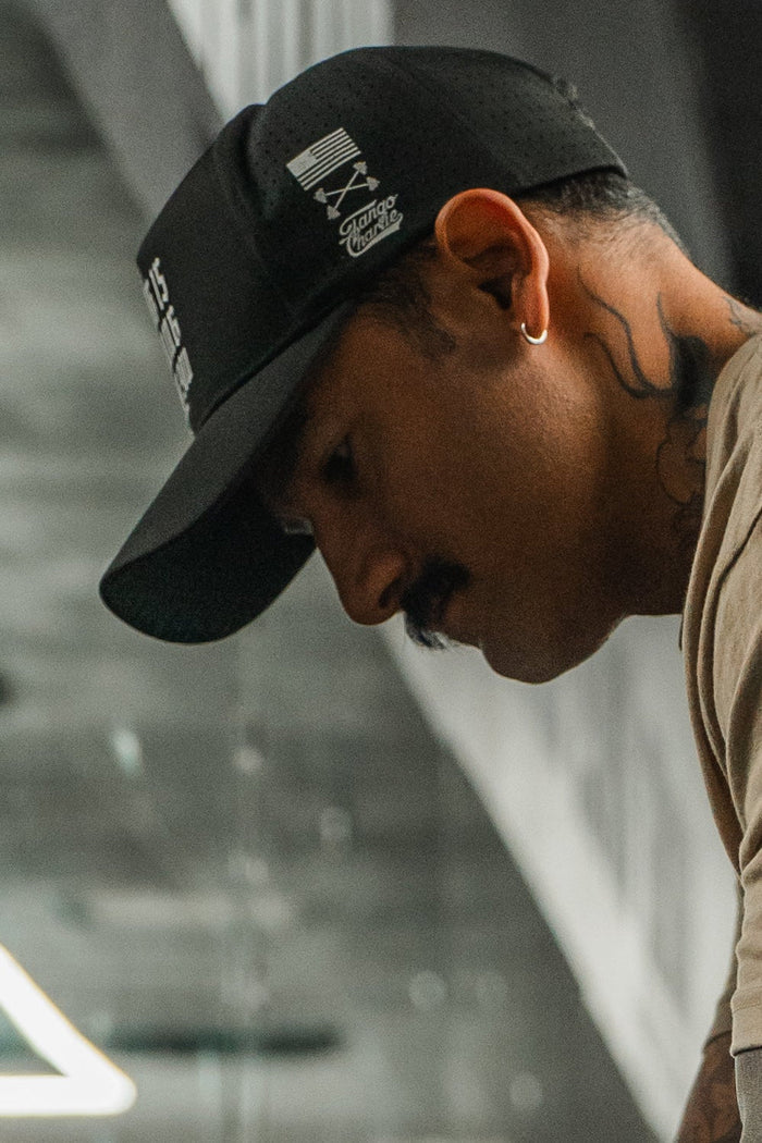 A man in the Tango Charlie Apparel "No One Is Coming To Save You Hydro" snapback hat, featuring black hydro-resistant fabric and white graphics, looks downward indoors. He wears a beige shirt, has tattoos on his neck, a mustache, and a hoop earring.