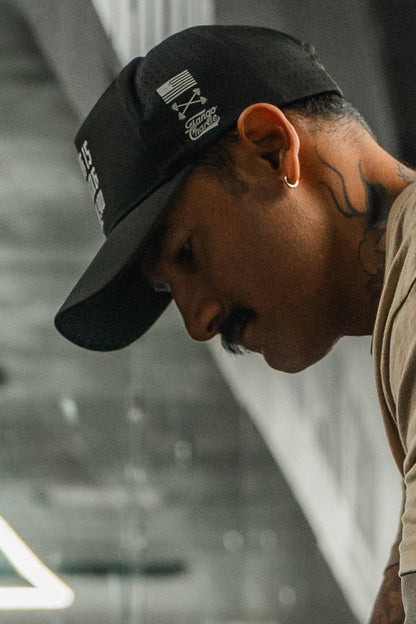 A man in the Tango Charlie Apparel "No One Is Coming To Save You Hydro" snapback hat, featuring black hydro-resistant fabric and white graphics, looks downward indoors. He wears a beige shirt, has tattoos on his neck, a mustache, and a hoop earring.