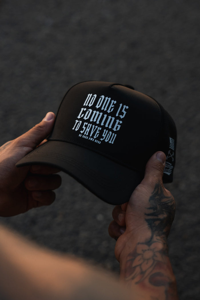 A tattooed arm holds the black No One Is Coming To Save You Hydro Snapback Hat by Tango Charlie Apparel, featuring bold white gothic text. A blurred dark background highlights the focus on the hat and hands.