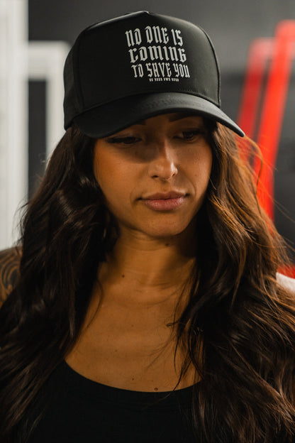A woman with long brown hair wears the Tango Charlie Apparel "No One Is Coming To Save You Hydro - Snapback Hat," a black hydro-resistant cap featuring the phrase in bold white letters. She is indoors, looking down with a neutral expression.