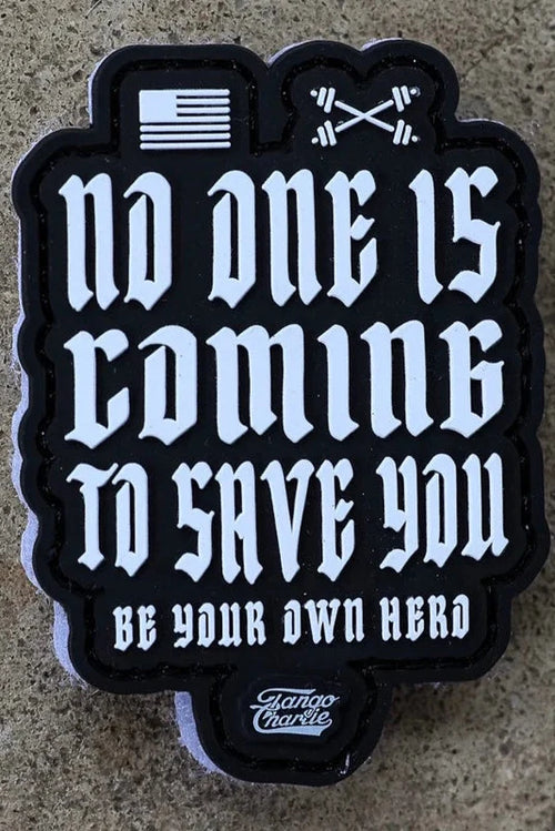 The No One Is Coming To Save You PVC Patch by Tango Charlie Apparel features white gothic text, a small flag, and crossed barbells above the message. The patch displays the Tango Charlie logo at the bottom and is shown on a stone surface.
