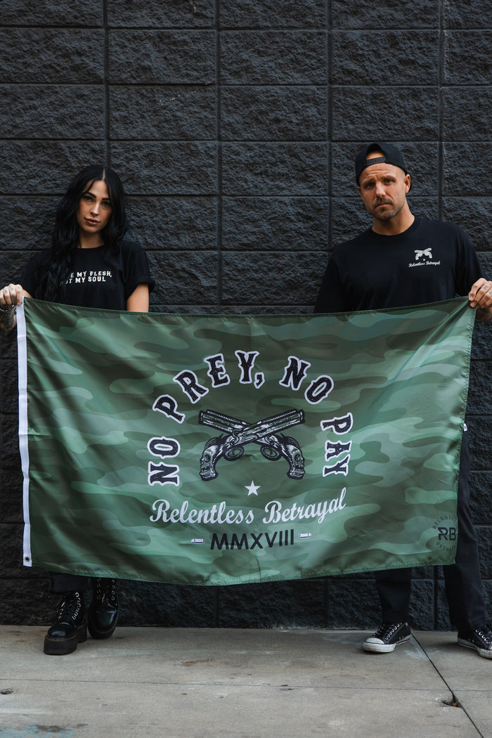 In front of a dark brick wall, two people hold the No Prey, No Pay Camo Flag by Relentless Betrayal, featuring crossed pistols and text about relentless betrayal. Their black attire and serious expressions perfectly match the rebellious spirit of the polyester flag displaying No Prey, No Pay.