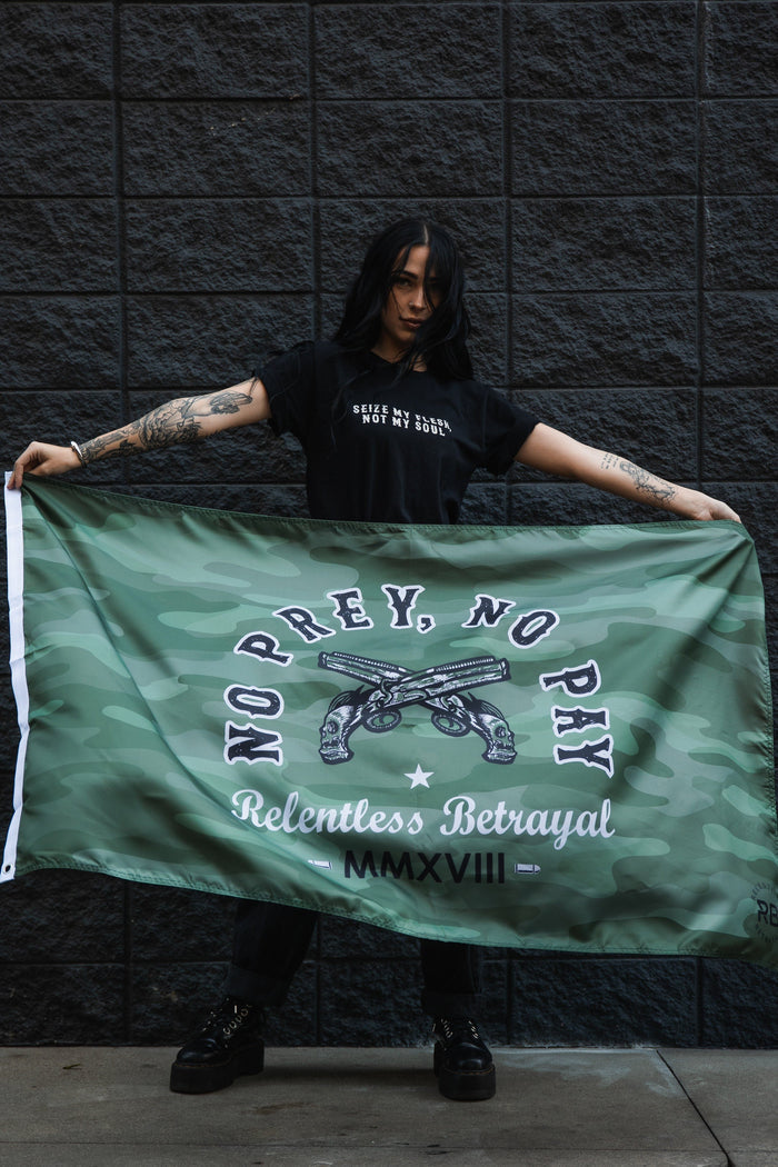 A tattooed person holds the No Prey, No Pay camo flag by Relentless Betrayal, made of durable polyester and featuring MMXVII. They stand against a dark brick wall in a black t-shirt with white text and matching pants.