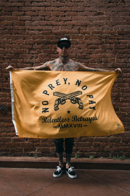 A person stands against a brick wall, holding the No Prey, No Pay Flag by Relentless Betrayal. The durable polyester flag is yellow, featuring crossed pistols with text: No Prey, No Pay and Relentless Betrayal MMXVIII. They wear tattoos, a cap, sunglasses, and casual attire.
