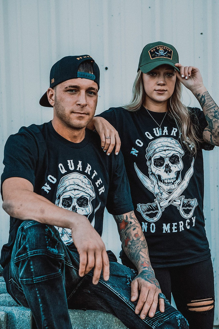 Two people wear matching No Quarter Black T-Shirts by Relentless Betrayal. The left person wears a black cap and classic tee, while the right person showcases tattoos and a green cap, embodying mercilessness against a light background.