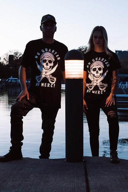 At dusk on a dock, a man and woman stand beside each other wearing Relentless Betrayals No Quarter Black T-Shirt. The shirts, featuring a skull and crossbones with No Quarter: No Mercy, are silhouetted by lamplight, adding an air of mercilessness to their figures against the water.