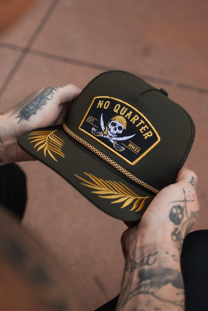 A tattooed individual holds a Relentless Betrayal No Quarter Olive Snapback, showing its skull design, gold embroidered leaves, and braided strap above the brim. The hat is water-resistant and olive in color.