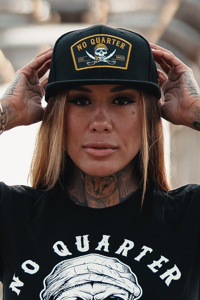 Sporting a No Quarter snapback from Relentless Betrayal, a person poses with one hand on the brim. The hat features a skull and crossbones design, complementing their tats on hands and neck, as they maintain a neutral expression.