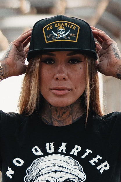 Sporting a No Quarter snapback from Relentless Betrayal, a person poses with one hand on the brim. The hat features a skull and crossbones design, complementing their tats on hands and neck, as they maintain a neutral expression.