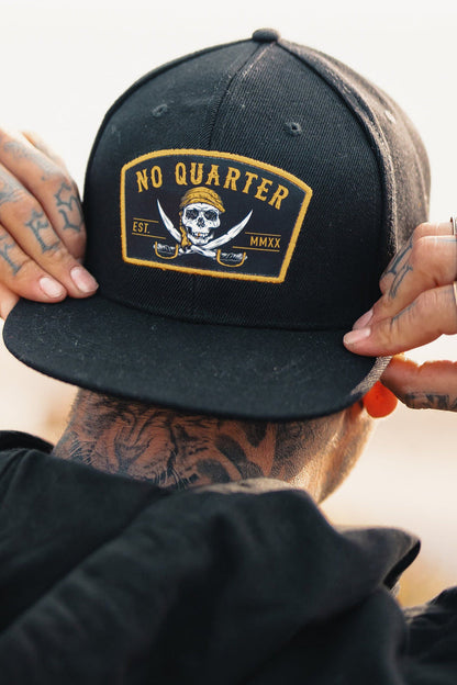 A person adjusts a Relentless Betrayal No Quarter Snapback featuring a skull and crossed swords with Est. MMXX. Tattoos adorn their neck and hands, complemented by an orange plug earring.