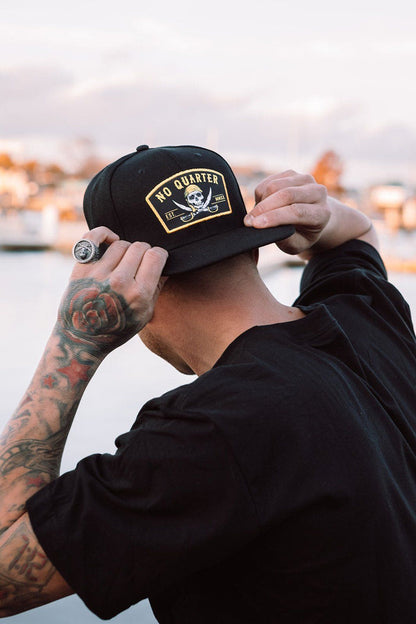 With tattooed arms, a person adjusts a Relentless Betrayal No Quarter Snapback featuring a skull and crossbones. They stand outdoors against a backdrop of blurred water and buildings.