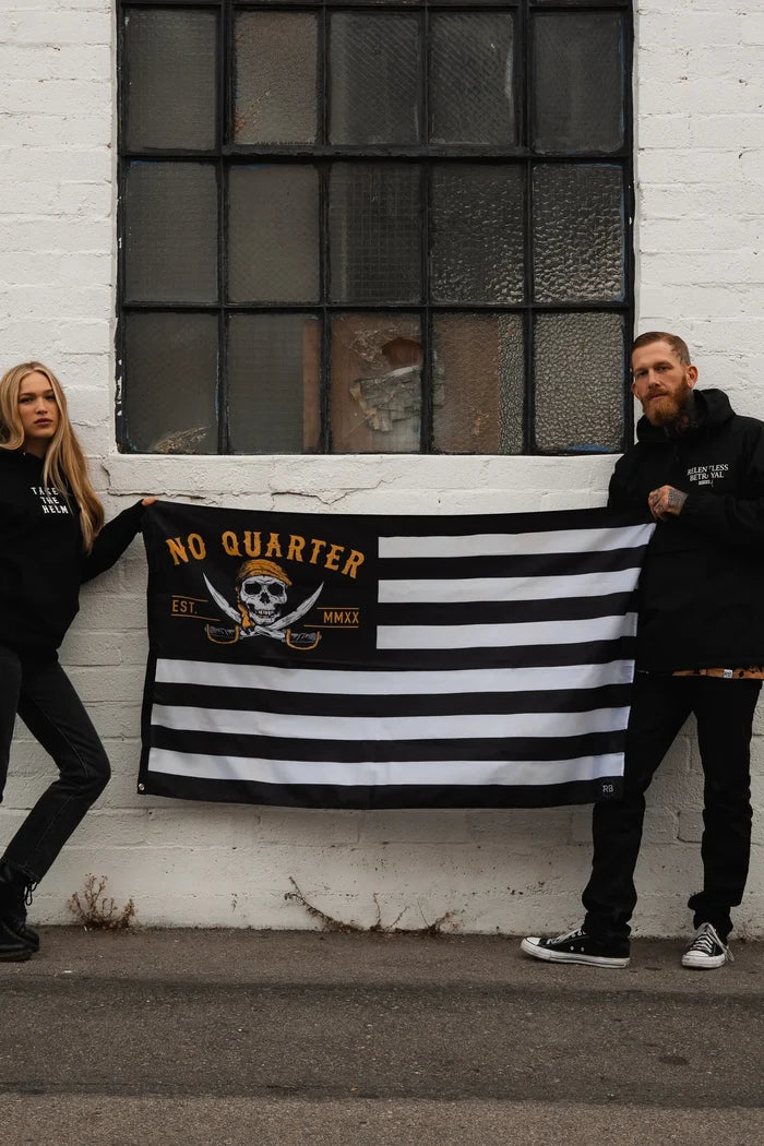 Two people stand against a white brick wall holding a durable No Quarter Stripe Flag V2 by Relentless Betrayal, featuring a skull and crossbones with No Quarter Est MMXX. The woman on the left leans casually, while the man on the right stands upright.