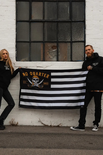 Two people stand against a white brick wall holding a durable No Quarter Stripe Flag V2 by Relentless Betrayal, featuring a skull and crossbones with No Quarter Est MMXX. The woman on the left leans casually, while the man on the right stands upright.