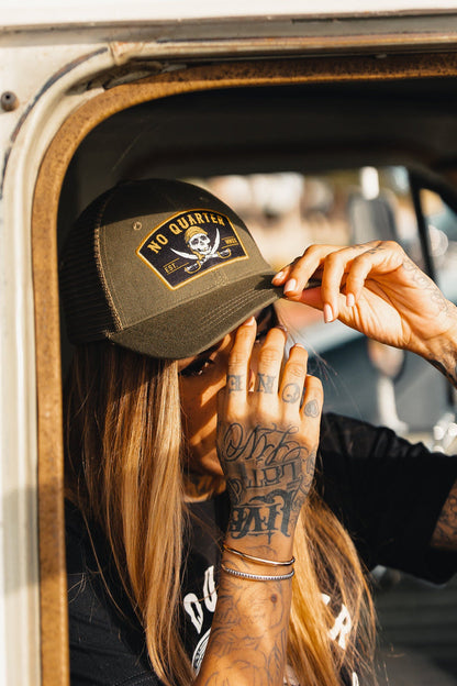 A person with long hair adjusts their Relentless Betrayal No Quarter Trucker Snapback, featuring a skull and crossbones. Leaning out the window, their intricately tattooed hand is visible.