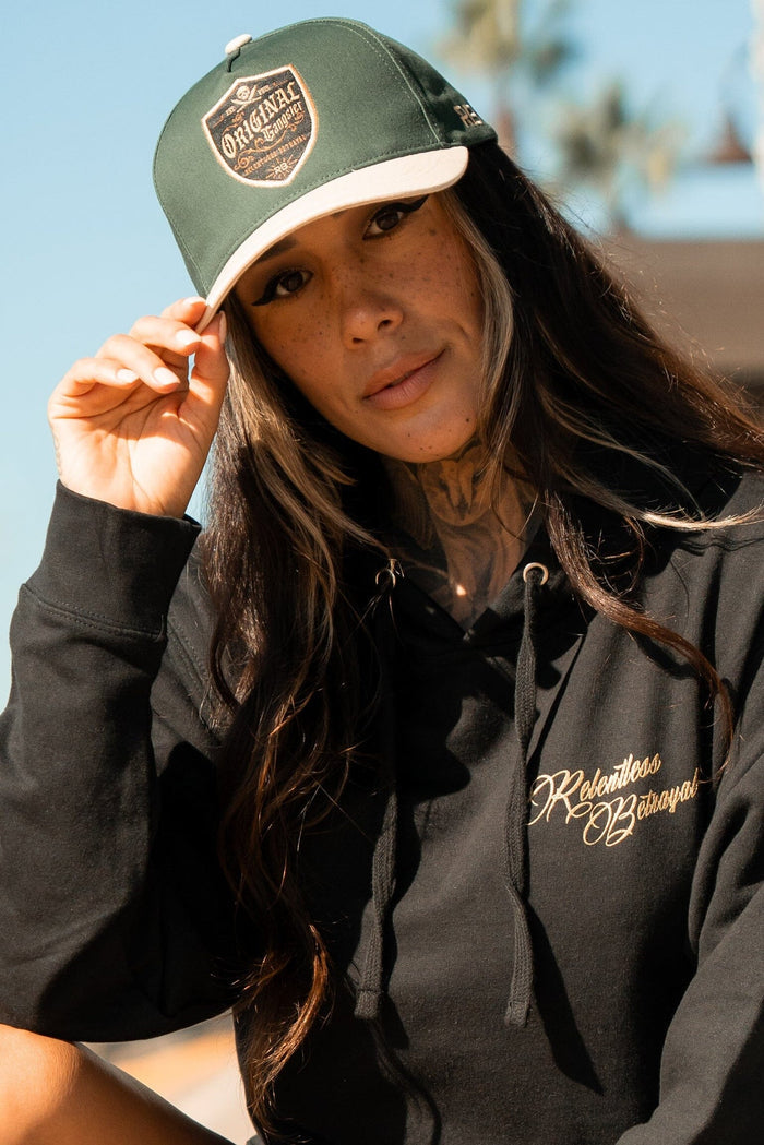 A person with long hair and tattoos wears a black hoodie emblazoned with Relentless Betrayal and an OG Forest Green Snapback. They adjust their cap outdoors against a backdrop of blurred trees and sky.