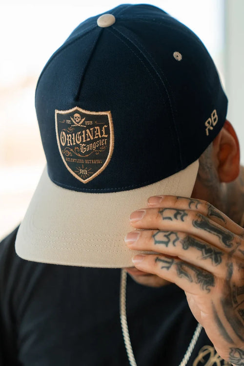 A tattooed person in a black T-shirt adjusts their OG Navy Snapback from Relentless Betrayal, featuring a patch labeled Original Gangster, a snapback enclosure, and the letters RB on the side.