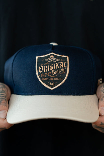 A man holds the OG Navy Snapback by Relentless Betrayal, featuring a shield-shaped patch with Original Gangster and intricate designs. Tattooed fingers grip the cap’s snapback enclosure tightly.