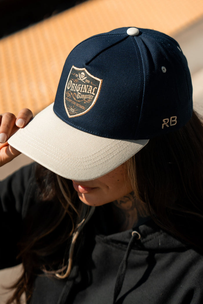 A person in a black hoodie adjusts their OG Navy Snapback from Relentless Betrayal, featuring a crest design that reads Original Gangster, a small RB on the side, and a Snapback Enclosure, while standing outdoors.