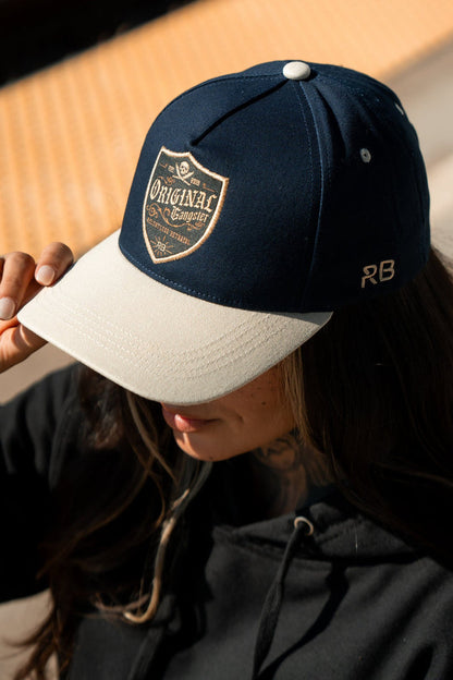 A person in a black hoodie adjusts their OG Navy Snapback from Relentless Betrayal, featuring a crest design that reads Original Gangster, a small RB on the side, and a Snapback Enclosure, while standing outdoors.