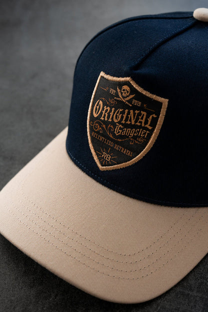 The OG Navy Snapback by Relentless Betrayal features a curved brim in navy and beige with a patch displaying Original Gangster, a skull motif, Est. 2015, and Relentless Betrayal within a shield, stylishly presented on a dark surface.