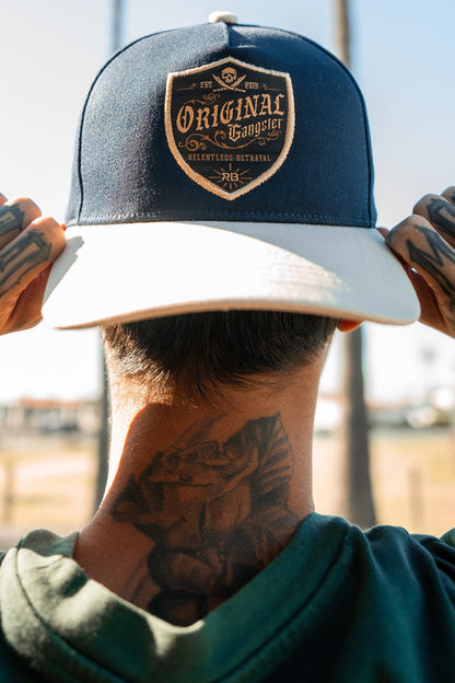 Amidst a sunny backdrop with palm trees, a person with tattooed hands and neck adjusts their OG Navy Snapback by Relentless Betrayal. The Original Gangster design adds an urban edge that complements their green shirt and enhances the scenes vibe.