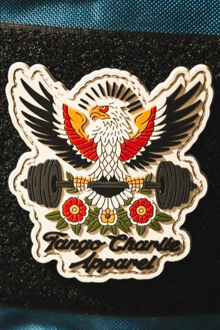 The “Old School, Never Gets Old - Patch” by Tango Charlie Apparel is a 3" 3D PVC patch showing an eagle with outstretched wings and a barbell, surrounded by flowers. The patch displays the brand name and is shown on black fabric.