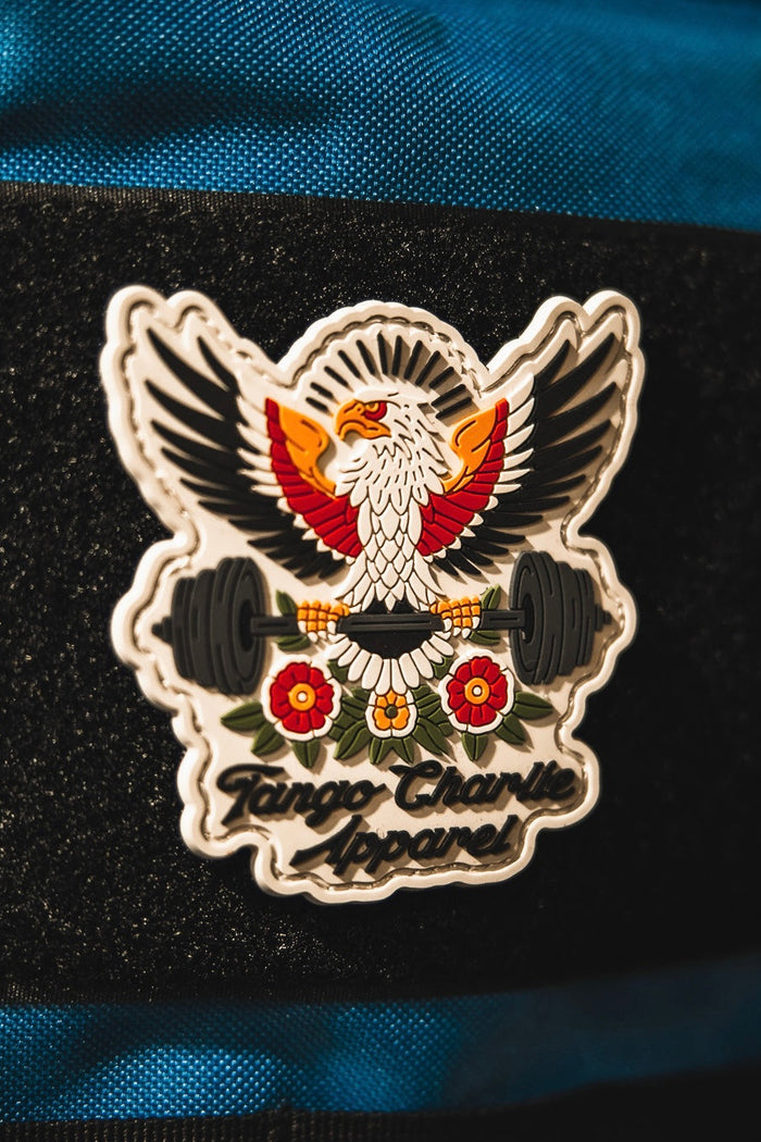 The "Old School, Never Gets Old - Patch" by Tango Charlie Apparel is a colorful 3D PVC patch showing an eagle with outstretched wings holding a barbell above two red flowers, attached to a blue fabric surface.