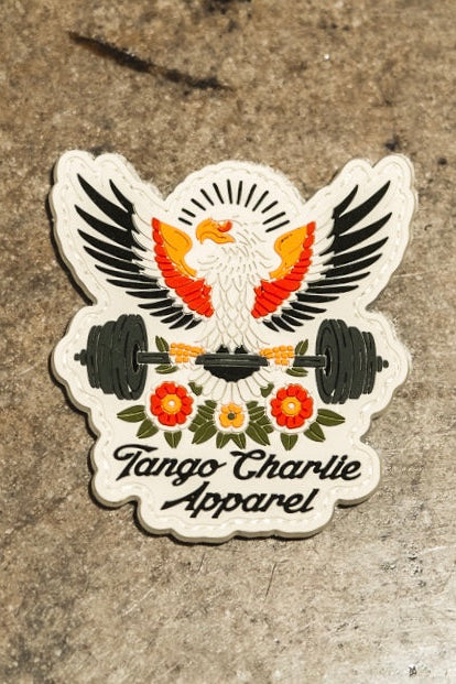 The "Old School, Never Gets Old - Patch" by Tango Charlie Apparel features an eagle with spread wings holding a barbell over red and yellow flowers, rays behind its head, and the brand name on a mottled grey background.