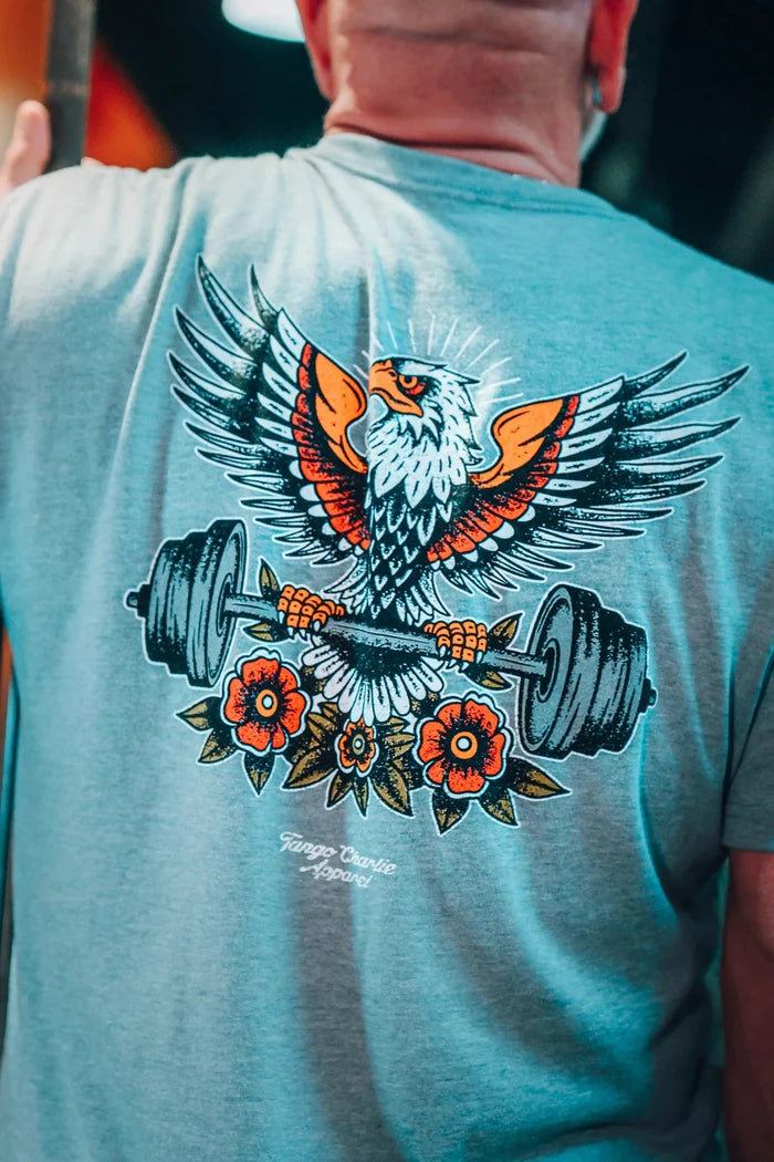A tattooed person wears the Tango Charlie Apparel Old School Never Gets Old gray shirt with a colorful eagle graphic, holding Cold Iron weightlifting plates with flowers below, capturing an old school gym vibe.