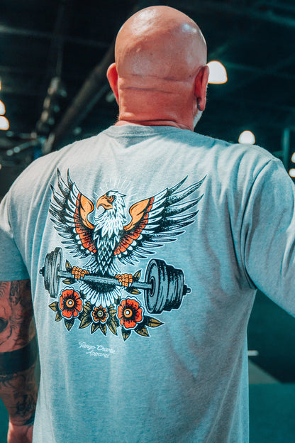 A bald, tattooed man wears Tango Charlie Apparel's "Old School Never Gets Old" gray tee with an illustrated eagle gripping weight plates and red flowers, set in a classic weightlifting gym.