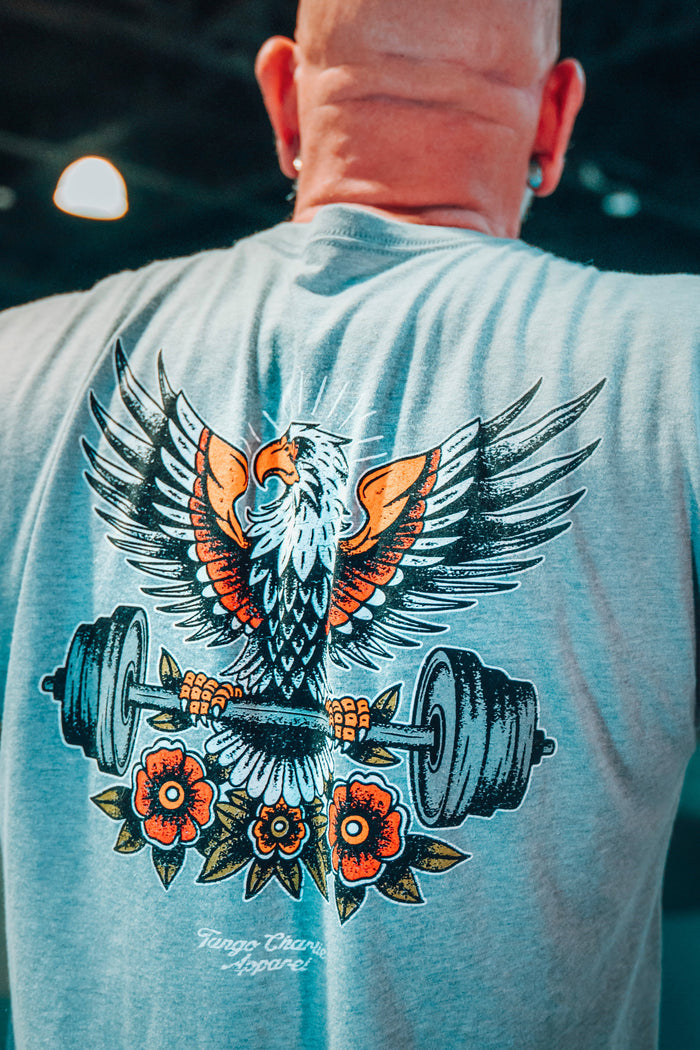 Person wearing a gray t-shirt with an eagle and barbell design on the back, standing in a gym.