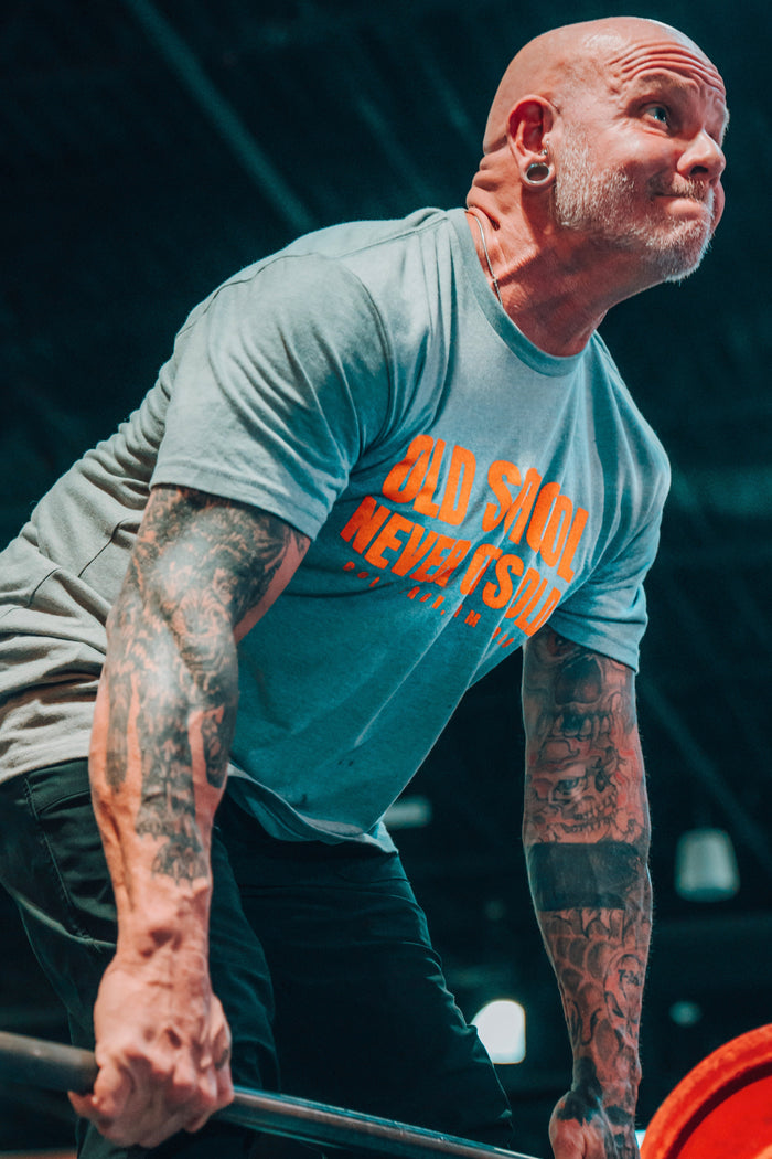 A muscular, tattooed man with a shaved head and large ear gauges lifts a barbell in an indoor gym, wearing Tango Charlie Apparel’s blue “Old School Never Gets Old” T-shirt. Classic weight plates are loaded on the bar.