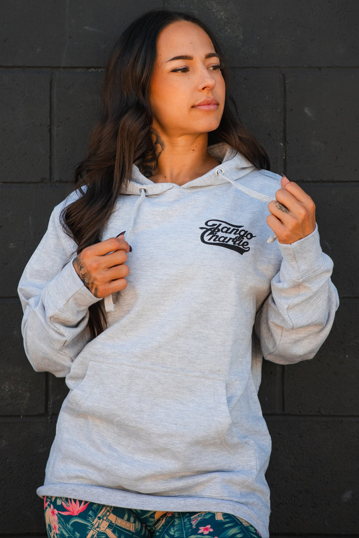A woman with long dark hair models the Only You Can Prevent Dad Bod Hoodie by Tango Charlie Apparel, standing before a dark brick wall and holding the hoodie strings while looking to the side.