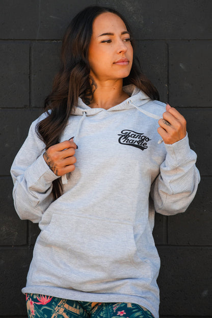 A woman with long dark hair models the Only You Can Prevent Dad Bod Hoodie by Tango Charlie Apparel, standing before a dark brick wall and holding the hoodie strings while looking to the side.