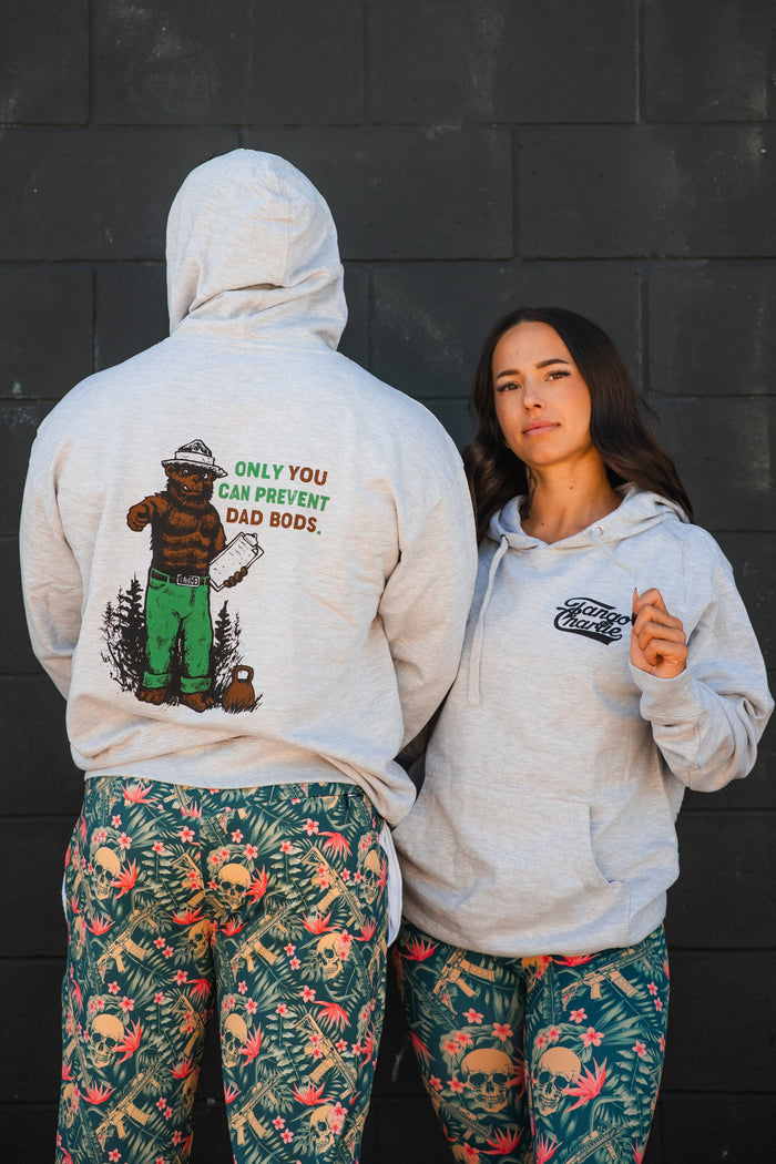 Two people in patterned leggings and gray Tango Charlie Apparel "Only You Can Prevent Dad Bod" hoodies stand by a dark wall. One shows the bear graphic with gym humor on the back; the other displays the front logo.