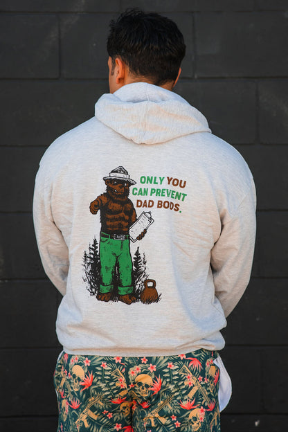 Wearing the Tango Charlie Apparel "Only You Can Prevent Dad Bod" Hoodie, a person faces away from a dark wall, pairing it with tropical gym humor shorts for a fun twist.