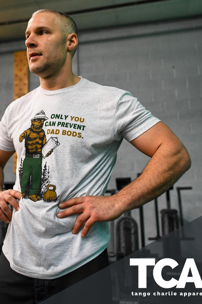 A person stands confidently in a gym wearing the Only You Can Prevent Dad Bod T-Shirt by Tango Charlie Apparel featuring a cartoon bear. Exercise equipment fills the background, with Tango Charlie Apparel and their logo displayed at the bottom right.