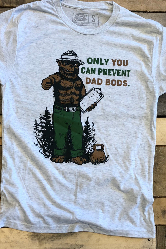 The Only You Can Prevent Dad Bod T-Shirt by Tango Charlie Apparel features a bear in green pants with a clipboard and the humorous slogan ONLY YOU CAN PREVENT DAD BODS, set against forest trees on a gray tri-blend fabric atop a wooden background.