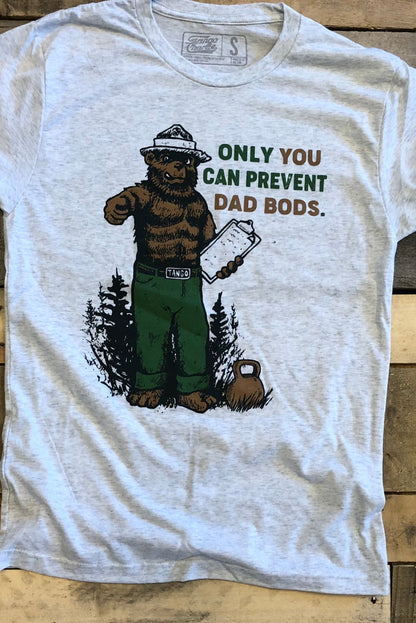 The Only You Can Prevent Dad Bod T-Shirt by Tango Charlie Apparel features a bear in green pants with a clipboard and the humorous slogan ONLY YOU CAN PREVENT DAD BODS, set against forest trees on a gray tri-blend fabric atop a wooden background.