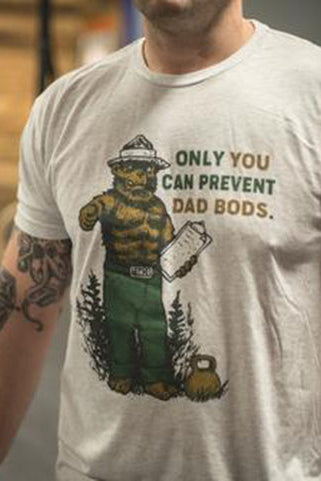 Wearing Tango Charlie Apparels Only You Can Prevent Dad Bod tri-blend T-shirt, a person showcases a whimsical bear in a ranger hat with clipboard. A tattoo graces their arm as they pose against a blurred indoor background.