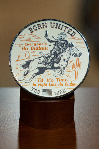The Outlaw Challenge Coin by Born United Private Label is a round metal coin featuring a cowboy on a bucking horse, two cacti, and the text: BORN UNITED. Everyone's an outlaw until it’s time to fight like an outlaw. TRD MRK.