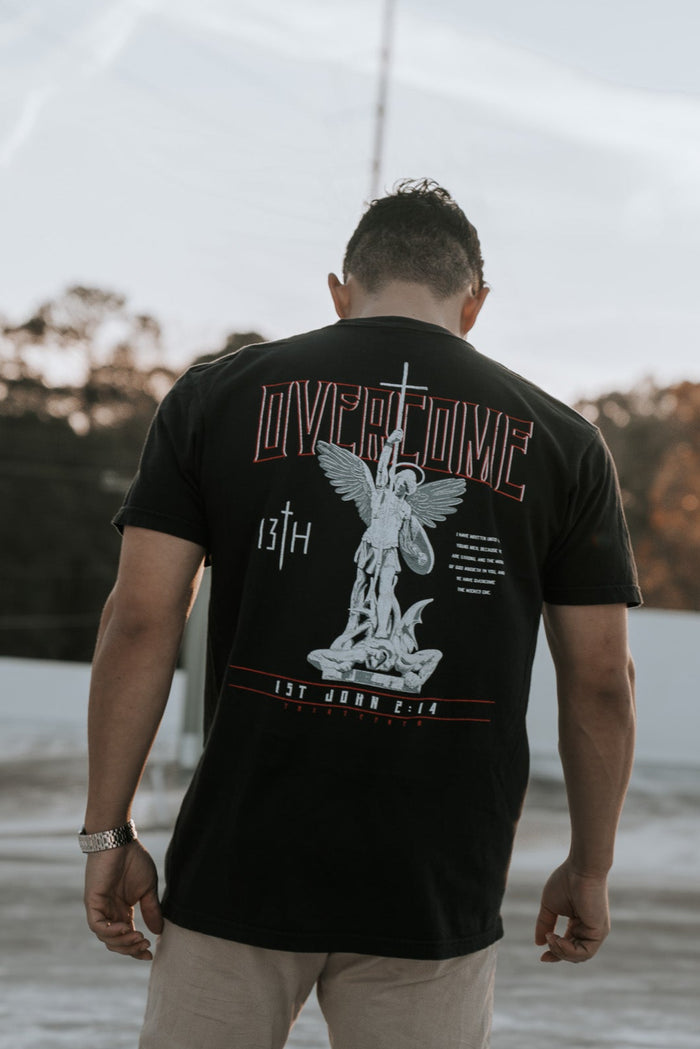 A young man stands outdoors with his back to the camera, wearing the Overcome Premium Tee by 13th. The black t-shirt features an angel graphic and Word of God, set against a serene backdrop of sunlit trees and light-colored ground.