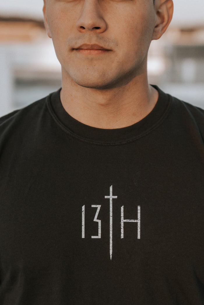A young man stands outdoors in the Overcome Premium Tee by 13th. With short dark hair and a neutral gaze, he looks into the distance, embodying resilience and the spirit to overcome. The background remains softly blurred.