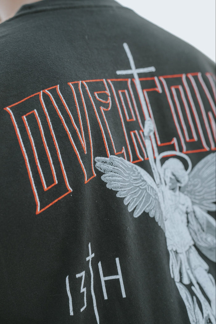 Close-up of the Overcome Premium Tee by 13th, a black shirt for young men. It features an angel graphic holding a spear and cross, with OVERCOME in red above and 13TH below, symbolizing strength through Gods word.